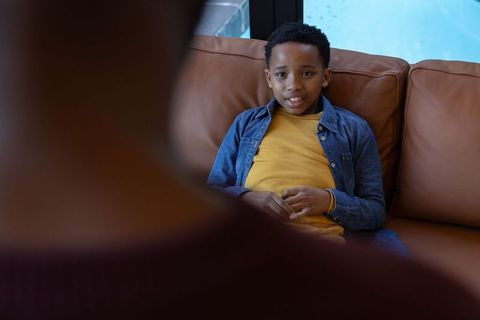 African american boy relaxing on leather sofa talking with caregiver by window