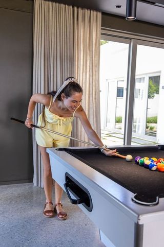 Young woman leaning and aiming cue on black-felt pool table in modern sunlit game room