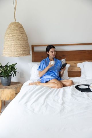 Woman relaxing in blue satin pajamas sipping coffee and scrolling smartphone on white bed