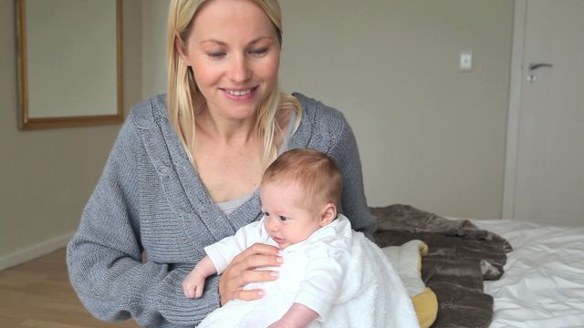 Woman smiling while playfully bouncing an infant on her knee in a cozy home setting. Ideal for parenting articles, imagery of motherhood and home life, or promotions of family products. It captures the tender bonding moment between a parent and child, demonstrating care and affection.