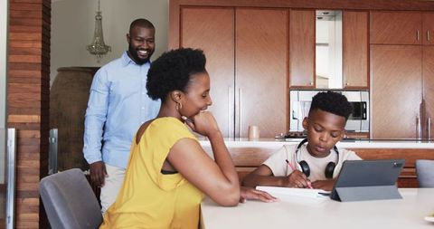 African american parents helping son studying at kitchen table with tablet and headphones