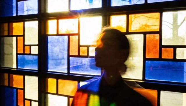 Standing silhouette facing stained-glass window catching colorful light through grid panes