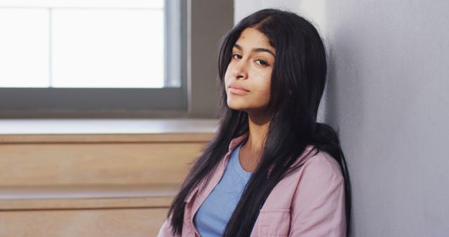 Young woman with long dark hair wearing pink jacket and blue shirt, leaning against wall indoors with serene, thoughtful expression. Natural light coming through window in the background. Ideal for content related to mental health, introspection, young adults, and peaceful moments.