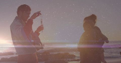 Parents checking phone while holding baby on rocky coast at dusk with lens flare