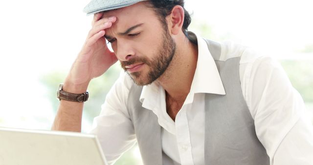Young man in white shirt, gray vest, and hat facing laptop with expression of concentration or frustration. Use for themes related to work stress, business challenges, modern office settings, freelance work, or professional contemplation.