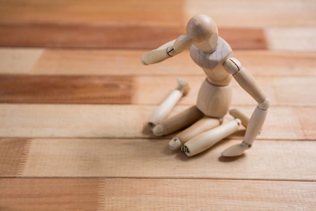 Wooden figurine sitting on wooden floor, depicting emotions of sadness and depression. Useful for illustrating concepts of mental health, loneliness, and emotional struggles. Ideal for articles, blogs, and educational materials on mental health awareness and emotional well-being.