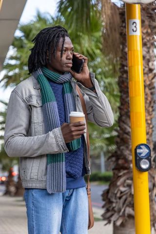 African american man talking on phone holding coffee at urban bus stop near palm trees