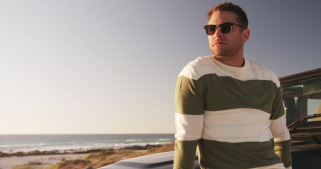Young man wearing sunglasses while leaning on a vehicle near the beachside during the day. Ideal for promoting travel destinations, summer vacation ideas, and casual fashion apparel. Useful for lifestyle blogs, coastal adventures, and outdoor activities.