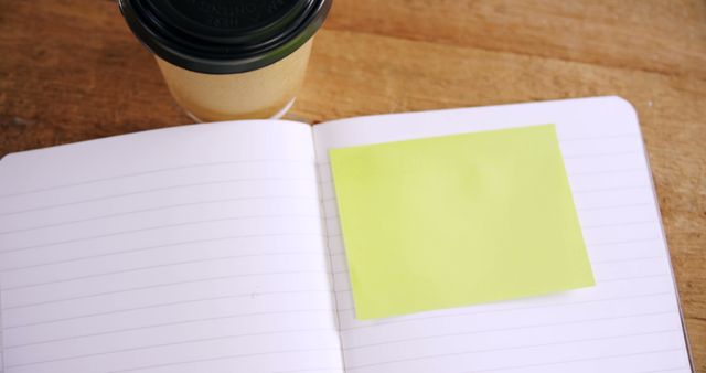 A blank notebook with a yellow sticky note lies open next to a takeaway coffee cup on a wooden surface, with copy space. Ideal for a message or reminder, the scene suggests productivity and planning.
