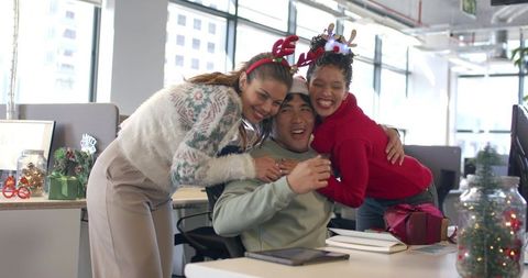 Diverse Coworkers Celebrating and Hugging Colleague Wearing Santa Hat at Office Desk with Gifts