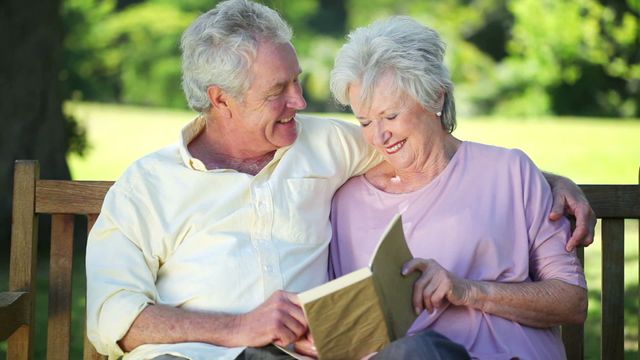 Senior couple sitting closely on a park bench under sunny skies. They are reading a book together, sharing smiles and displaying affection. Ideal for use in advertisements related to elderly care, retirement plans, family bonding, leisure and lifestyle campaigns promoting senior well-being.