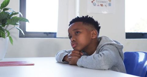 Pensive student resting chin on desk in bright classroom wearing gray hoodie and blue chair