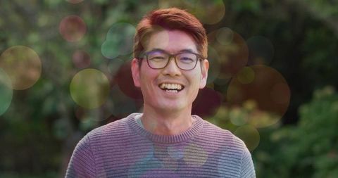 Smiling korean man wearing round glasses standing in garden with warm bokeh light
