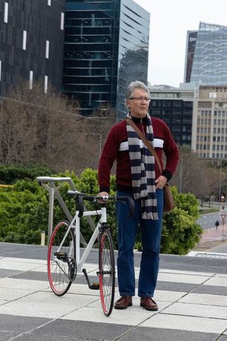 Senior commuter standing with bicycle on urban plaza wearing striped scarf and satchel