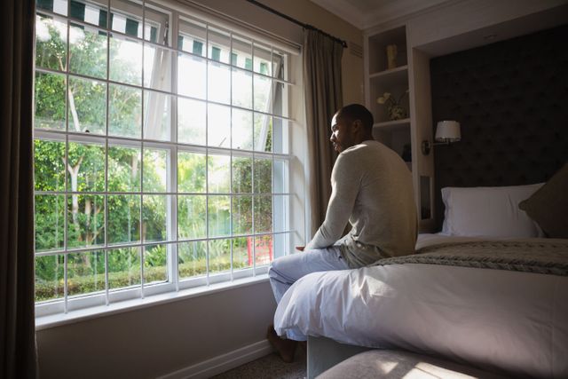 Man sitting on bed by window in cozy bedroom, looking outside. Ideal for themes of relaxation, contemplation, morning routines, and home life. Perfect for use in articles, blogs, or advertisements related to mental health, lifestyle, interior design, and personal well-being.