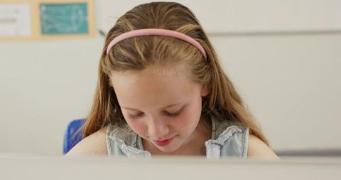 Preteen girl focusing on book at classroom desk wearing pink headband and denim top