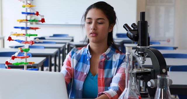 Female student in a science classroom studying a DNA model and using a laptop. Next to her is a microscope and laboratory equipment, suggesting engagement in scientific research or a school project. Perfect for content related to education, STEM fields, student life, and academic resources.