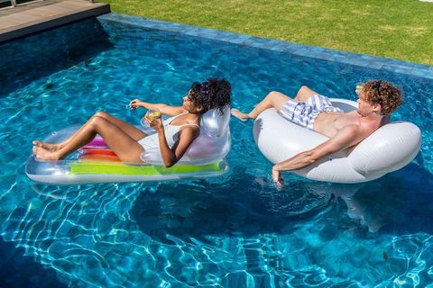 Couple lounging on inflatable pool floats drinking cocktails in sunny backyard pool
