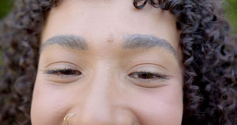 a close-up view of a woman with natural curly hair expressing subtle joy. she is adorned with a fashionable nose ring, adding to her elegant style. this visual can be ideal for use in beauty and fashion magazines, blogs highlighting personal expressions, or materials focused on self-confidence and modern trends.