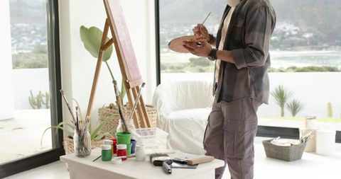 Indian male artist painting at easel in sunlit waterfront studio with palette and brushes