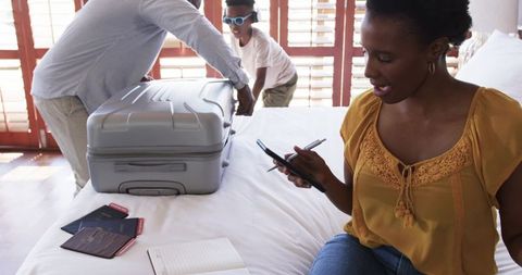 African american family packing luggage in sunlit hotel room using phone and passports