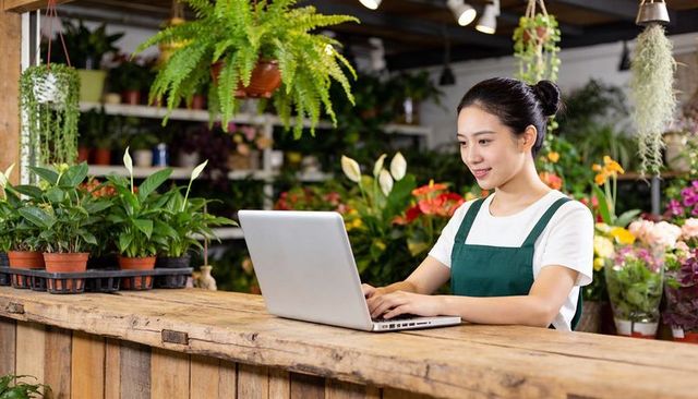 Young florist managing online orders at rustic plant shop counter using laptop
