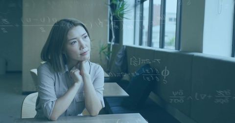 Gazing asian woman resting chin on clasped hands at wooden table with formulas overlay
