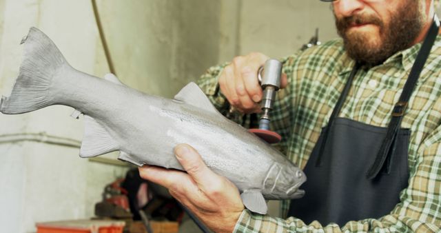A middle-aged Caucasian man is meticulously working on a fish sculpture, with copy space. His focus and craftsmanship suggest he is an artist or sculptor in his workshop.