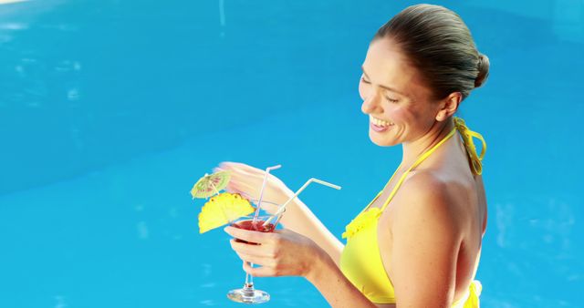 Depicts a happy woman in a yellow swimsuit standing in a pool holding a tropical drink with a smile. Perfect for use in travel advertisements, summer holiday promotions, wellness and lifestyle blogs, social media posts, or beverage commercials.