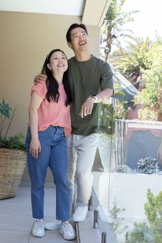 Asian couple standing on sunlit balcony smiling and looking up by glass railing