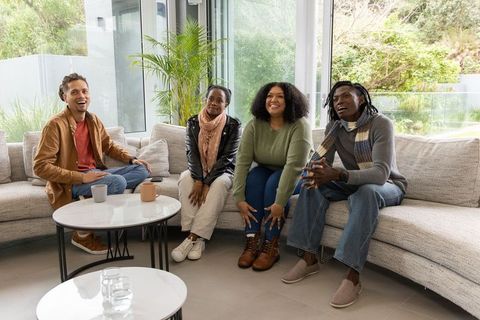 Diverse friends laughing and chatting on curved sofa holding mugs in modern living room