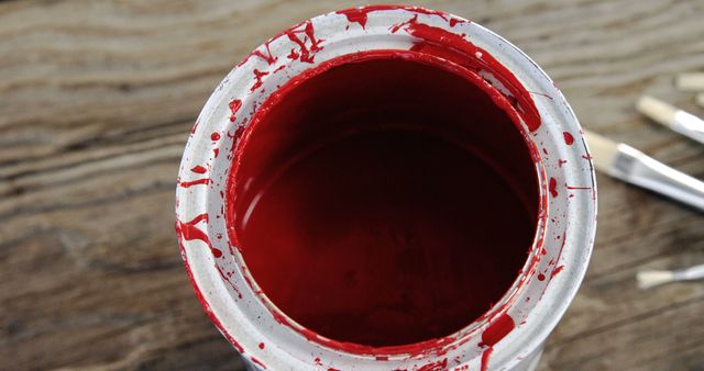 Top view of open can of red paint placed on a rustic wooden surface with some paint brushes in the background. The close up captures splatters and smudges, highlighting the idea of a work in progress. Perfect image for topics related to DIY projects, home improvement, art and crafts, decorative ideas, and creative endeavors.