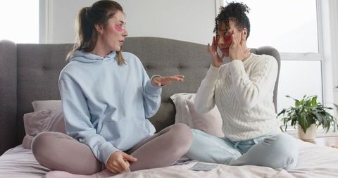 Friends applying under-eye patches on bed during cozy at-home skincare session