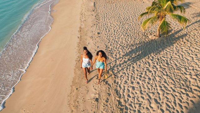 Two women walking hand in hand along tropical beach barefoot with palm tree and footprints