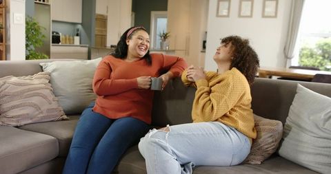Two women laughing on cozy sectional sofa sharing coffee and relaxed conversation