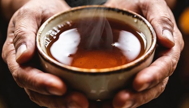 Senior hands cradling steaming amber tea in rustic ceramic cup, closeup warm comfort