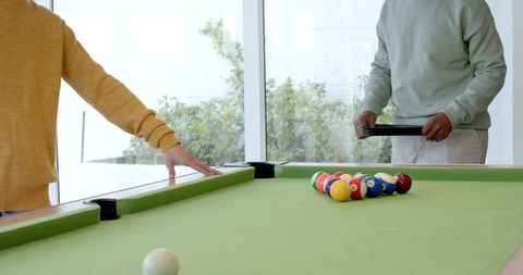 Diverse friends racking colorful billiard balls on sunlit green pool table by windows