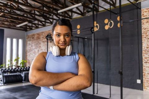 Confident indian woman standing arms crossed in gym wearing headphones and blue workout top