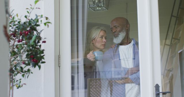 Worried senior diverse couple in kitchen, looking through window and embracing. retirement lifestyle, spending time at home.