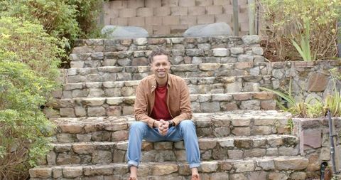 Smiling man sitting on rustic stone steps in backyard garden wearing brown jacket