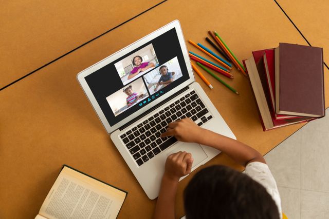Kid is engaged in online class, interacting with teacher and classmates through video call on laptop. Ideal for illustrating digital education, remote learning environments, and online school resources. Background includes pencils and educational books, enhancing academic theme.