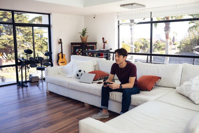 Asian teenage boy sitting on a white couch in a modern living room, focused on playing a video game. The room features musical instruments, large windows, and a bright, airy atmosphere. Ideal for use in articles or advertisements related to gaming, technology, teenage lifestyle, home entertainment, or modern living spaces.