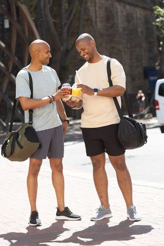 Clinking post-workout drinks while carrying gym bags on sunny urban sidewalk