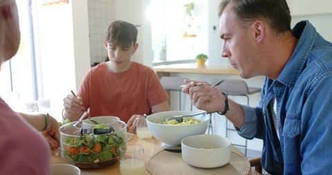 Family sharing casual healthy lunch at kitchen table with fresh salad, pasta and juice