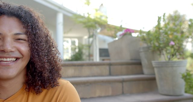 Young man with curly hair on steps in garden, smiling, enjoying outdoors on a sunny day. Suitable for use in lifestyle, happiness, and community-focused projects.