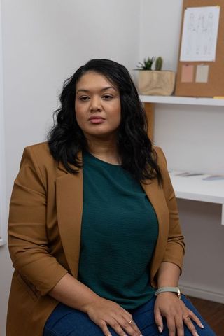 Thoughtful Asian Businesswoman in Tan Blazer Sitting in Minimal Studio with Corkboard Sketches