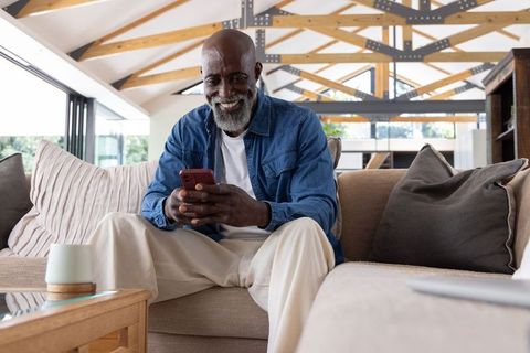 Senior african american man smiling while using smartphone on beige sofa in sunlit loft