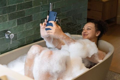 African american woman taking selfie in bubble bath, relaxing in green-tiled bathroom