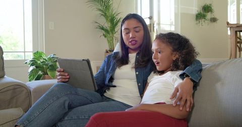 Diverse mother and daughter relaxing on sofa sharing tablet, smiling and bonding in cozy living room