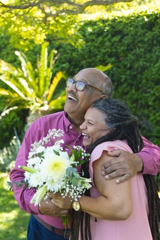 Senior couple embracing and laughing in sunlit garden with bouquet of white flowers